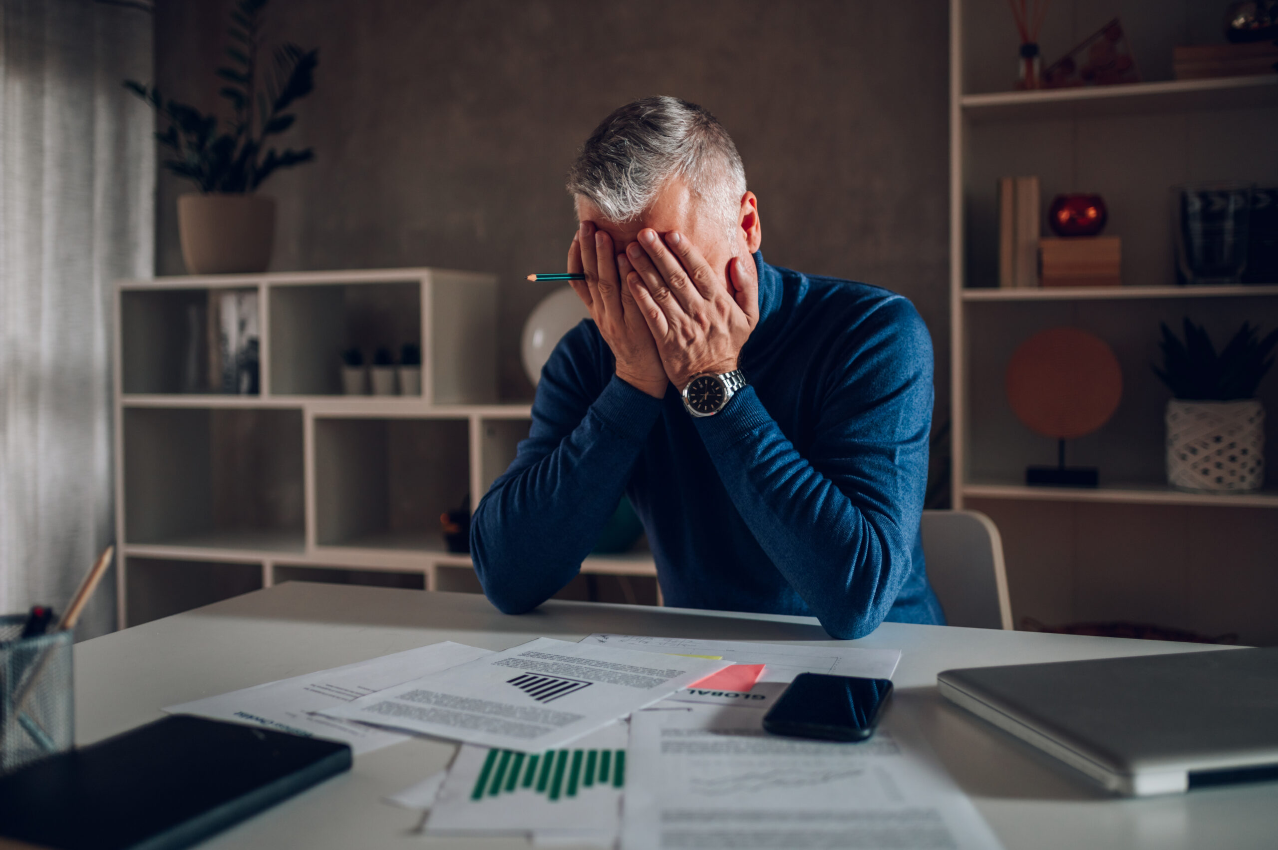 Middle aged man working at home at modern workplace and feeling stressed and tired. Businessman with gray beard and hair covering his face with hands. Working remotely in a modern work station at home